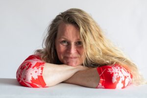 Wilma de Boer met lang blond haar in rood met witte jurk, leunend met armen op tafel en kijkend naar de camera